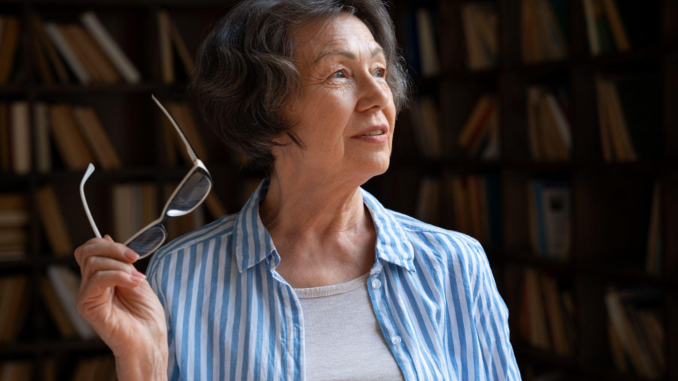 Esta imagem apresenta uma mulher madura em um ambiente que sugere uma biblioteca ou escritório pessoal.