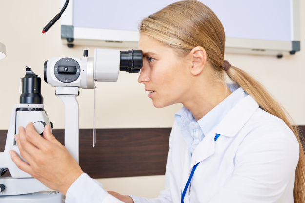 médica loira usando jaleco branco realizando exame oftalmológico dentro de seu consultório