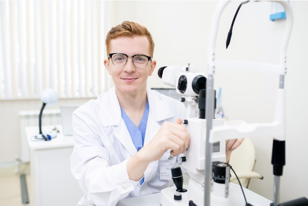 oftalmolista ruivo usando óculos e jaleco branco em seu consultório preparando seu equipamento para realizar retinografia simples