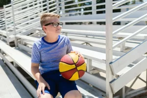 Menino de óculos segurando bola de basquete sentado em uma arquibancada de madeira