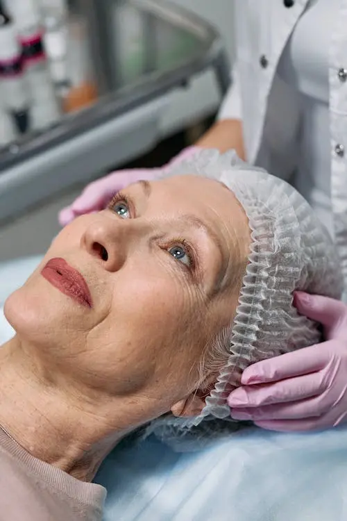 Mulher idosa de olhos azuis deitada com uma toca de cirurgia e mãos de luva rosa segurando sua cabeça