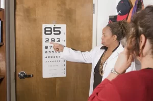 Mulher de blusa vermelha fazendo exame de vista com uma mulher negra de jalkeco apontando para um painel com números de diferentes tamanhos