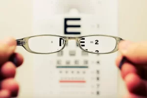 Mãos segurando óculos de grau com letras de teste oftalmologico ao fundo, embaçado ao redor do óculos
