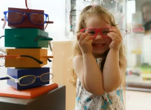 Menina usando óculos vermelho, seguro os com as mãoes, ao lado de um bloco colorido com óculos de grau expostos
