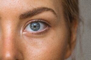 Imagem close-up de um olho humano. O olho está em foco, mostrando a íris azul e a pupila preta no centro. Há uma leve reflexão de luz na superfície da córnea, e a pele ao redor do olho é visível