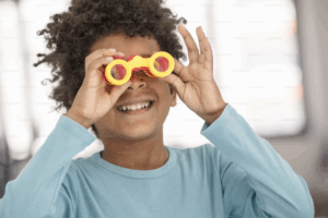 A imagem mostra uma criança sorridente usando um binóculo de brinquedo, simbolizando curiosidade e interesse visual. A cena representa a importância de estimular e observar a saúde dos olhos de forma lúdica, especialmente no contexto do retorno às aulas.
