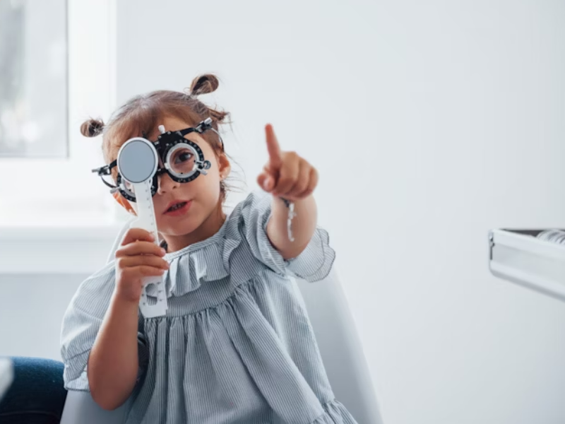 A imagem mostra uma criança durante um exame oftalmológico. Ela está usando um aparelho de teste de visão e segurando um oclusor ocular, enquanto aponta para algo à frente — um possível estímulo visual do teste. A cena é clara, bem iluminada e transmite um ambiente clínico acolhedor.