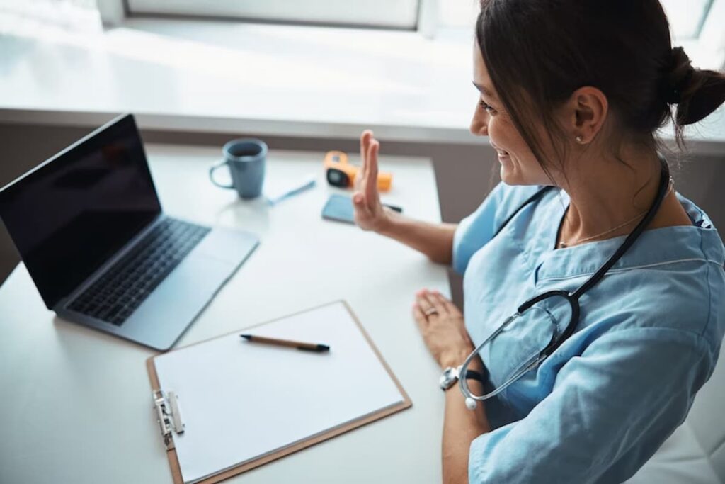 Na imagem que você enviou, vemos uma profissional da saúde — provavelmente uma médica ou enfermeira — usando uniforme e estetoscópio, interagindo com alguém por meio de uma videochamada em um notebook.