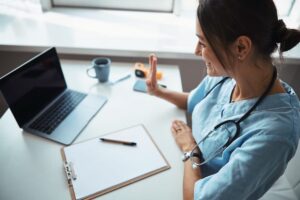 Na imagem que você enviou, vemos uma profissional da saúde — provavelmente uma médica ou enfermeira — usando uniforme e estetoscópio, interagindo com alguém por meio de uma videochamada em um notebook.