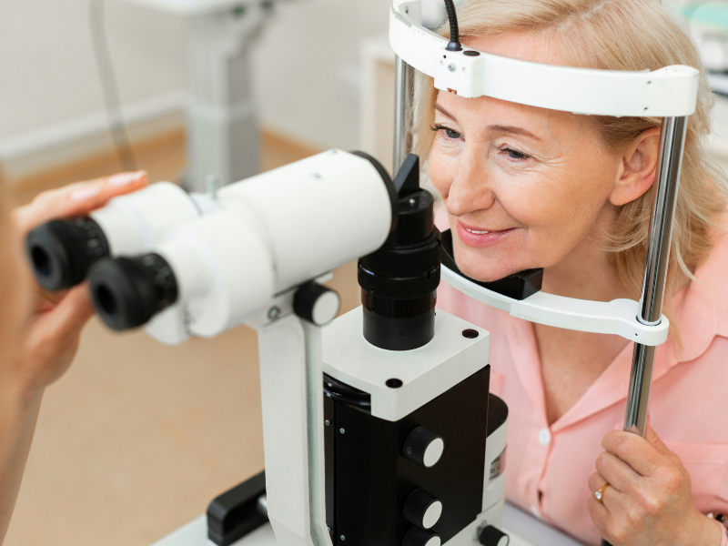 A imagem mostra uma mulher idosa realizando um exame oftalmológico com lâmpada de fenda (biomicroscópio), um equipamento utilizado por oftalmologistas para examinar com precisão as estruturas dos olhos, como córnea, íris, cristalino e retina.