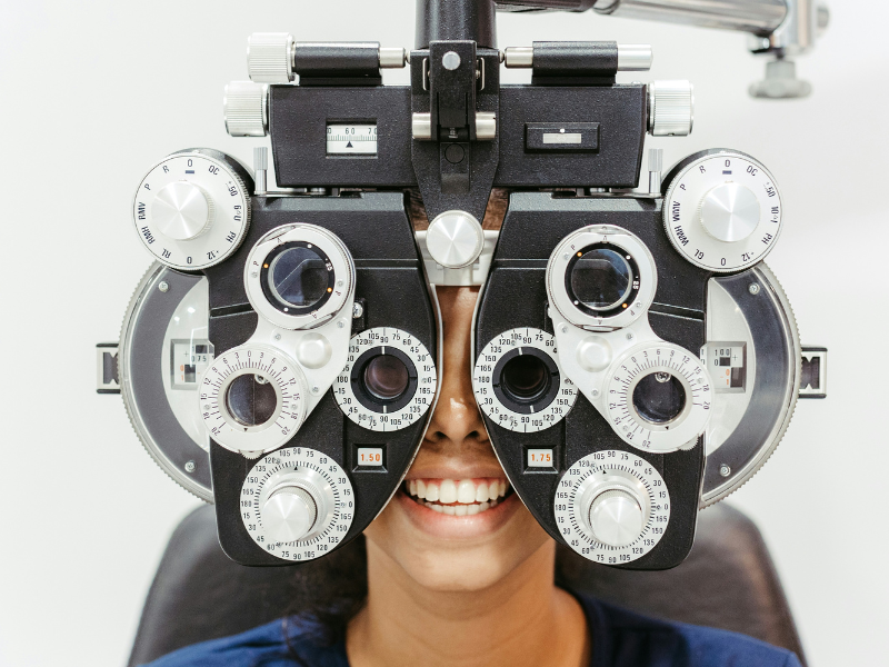 A imagem mostra uma pessoa sorridente realizando um exame oftalmológico com o uso de um foróptero, um instrumento utilizado para medir o grau de refração dos olhos. O foróptero está posicionado à frente do rosto da pessoa, cobrindo seus olhos, enquanto diferentes lentes são testadas para avaliar a acuidade visual e determinar a prescrição de óculos ou lentes de contato.
