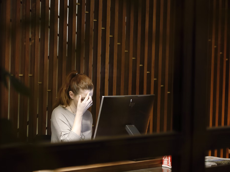A imagem mostra uma mulher sentada em frente a um monitor de computador, com a mão no rosto, especificamente cobrindo os olhos, em um gesto que sugere fadiga visual, estresse ou cansaço mental. O ambiente parece ser um escritório ou recepção, com divisórias de madeira ao fundo e iluminação quente. A mulher tem cabelos castanhos claros ou ruivos, presos em um rabo de cavalo, e veste uma blusa cinza-clara.