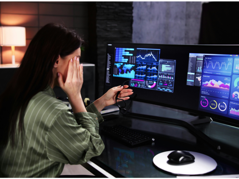 A imagem mostra uma mulher sentada em frente a um monitor ultra-wide, exibindo vários gráficos e dashboards de dados. Ela segura seus óculos com uma mão e esfrega os olhos com a outra, sinalizando claramente fadiga ocular ou cansaço visual após um longo período de trabalho em frente à tela.
