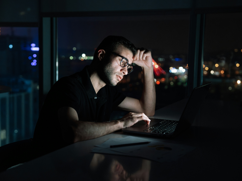 A única luz visível na cena vem da tela do computador, que ilumina parcialmente o rosto do homem e parte da mesa à sua frente. Ele parece cansado ou concentrado, apoiando a cabeça com uma das mãos enquanto digita com a outra.
