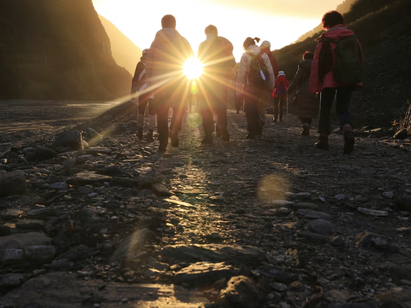 A imagem mostra um grupo de pessoas caminhando por uma trilha de terra e pedras em meio à natureza, durante o nascer ou pôr do sol. O sol está posicionado diretamente à frente do grupo, criando um forte brilho dourado e feixes de luz que irradiam para a lente da câmera.