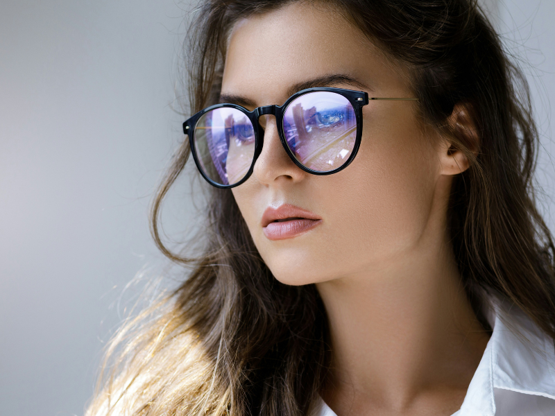 A imagem mostra uma mulher jovem com expressão neutra, usando óculos com lentes que possuem reflexo azul-violeta, típico de lentes com filtro de luz azul. Esse tipo de lente é projetado para proteger os olhos da exposição prolongada à luz azul emitida por telas de computadores, celulares e outros dispositivos eletrônicos.