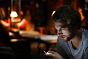 A imagem mostra um homem jovem com barba e cabelo claro, sentado em um ambiente escuro e acolhedor, iluminado principalmente pela luz do celular que ele segura nas mãos. Ele está olhando atentamente para a tela do aparelho, o que ilumina seu rosto de forma intensa, destacando suas feições contra o fundo escuro.