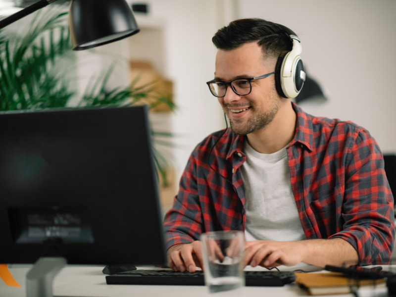 A imagem mostra um homem jovem sorrindo enquanto trabalha em frente a um computador de mesa. Ele está usando óculos com lentes transparentes (possivelmente com filtro de luz azul) e um fone de ouvido grande cobrindo as orelhas, sugerindo que está em uma videoconferência, reunião online ou jogando.