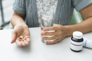 A imagem mostra uma pessoa prestes a tomar duas cápsulas com um copo de água, ao lado de frascos de suplementos. Ela transmite a ideia de cuidado diário com a saúde, especialmente ligada à prevenção de problemas visuais.