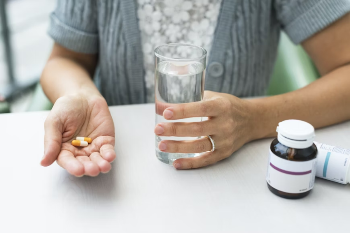A imagem mostra uma pessoa prestes a tomar duas cápsulas com um copo de água, ao lado de frascos de suplementos. Ela transmite a ideia de cuidado diário com a saúde, especialmente ligada à prevenção de problemas visuais.