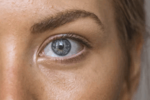 A imagem é um close-up de um olho. O olho tem uma íris de cor azul clara. A pupila, que é preta, reflete uma luz branca. Ao redor do olho, é possível ver a pele, algumas rugas finas e alguns pelos das sobrancelhas. A imagem está nítida e bem focada no olho.