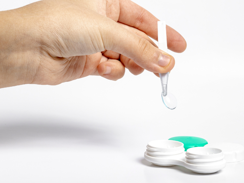 A imagem mostra uma mão segurando uma pinça de silicone com uma lente de contato gelatinosa transparente, prestes a ser colocada ou retirada de um estojo apropriado para lentes de contato. O fundo é branco, reforçando o aspecto limpo e estéril da cena.