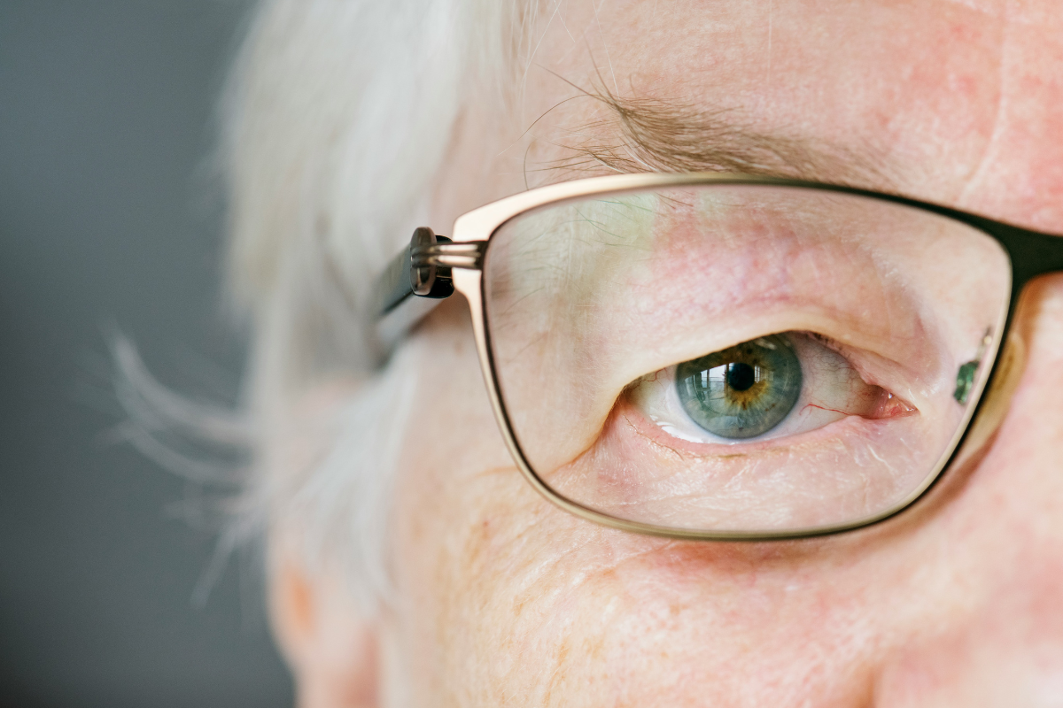 A imagem é um close-up do olho de uma pessoa idosa, possivelmente uma mulher, com cabelo branco. O foco está no seu olho esquerdo, que tem a íris em um tom de azul esverdeado com um leve anel cinza na borda