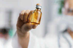 A imagem mostra uma pessoa usando jaleco e máscara segurando um pequeno frasco de vidro transparente com tampa metálica, contendo um líquido amarelo claro, possivelmente metanol.