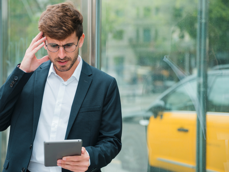 A imagem retrata um homem jovem, vestido de forma profissional, com uma expressão de preocupação, frustração ou dor de cabeça, enquanto interage com um tablet.