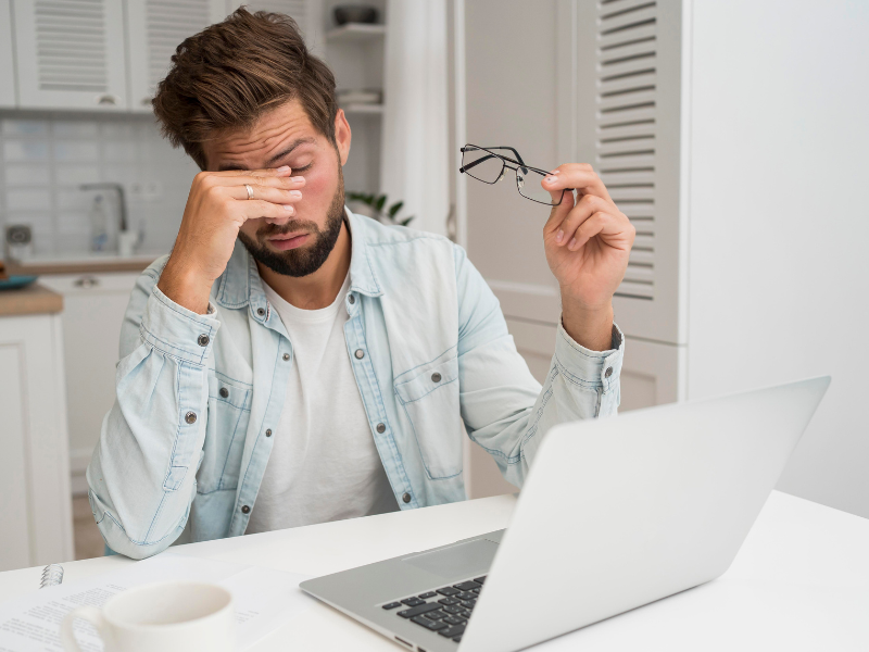 A imagem mostra um homem jovem com barba, sentado em uma mesa e visivelmente desconfortável com seus olhos, o que sugere fadiga visual ou a "Síndrome da Visão do Computador"