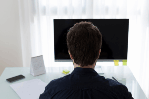 A imagem mostra a parte de trás da cabeça e ombros de um homem sentado em uma mesa de escritório, olhando diretamente para um monitor de computador.