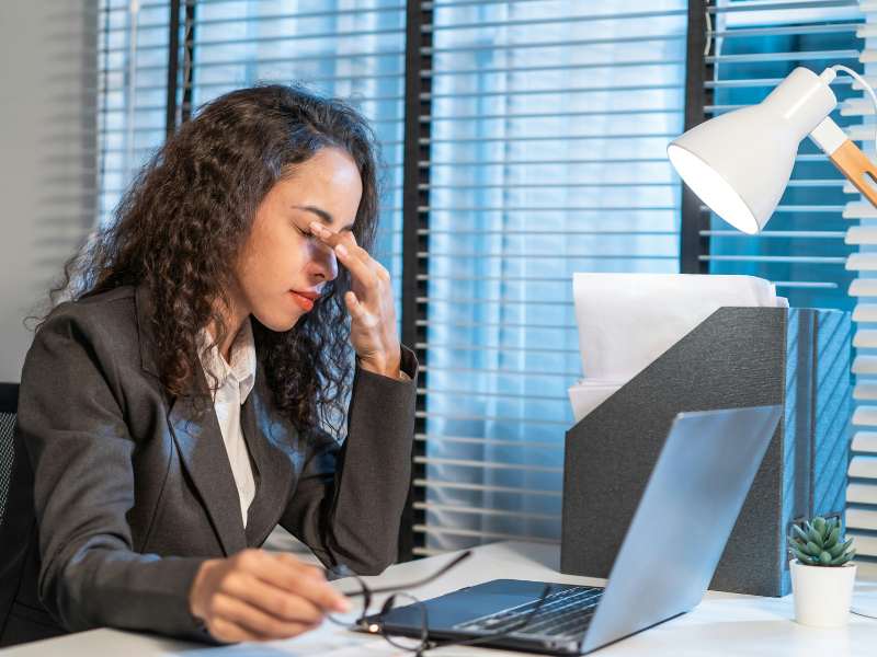 A imagem retrata uma mulher de negócios ou profissional em seu local de trabalho, expressando cansaço visual.