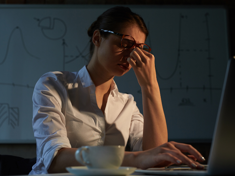 A imagem mostra uma mulher em um ambiente de escritório ou de trabalho, sob luz baixa (noturna ou ambiente escuro) e dor nos olhos
