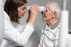A imagem é uma fotografia em close-up que retrata um exame oftalmológico ou de vista realizado por uma profissional de saúde em um paciente idoso.