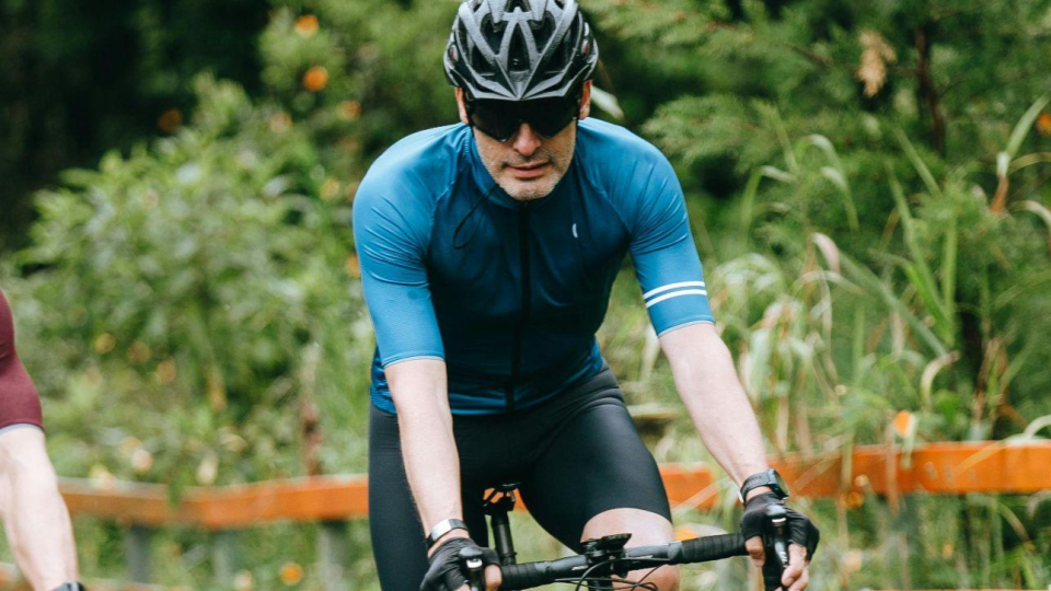 Ele usa capacete preto, óculos escuros esportivos e luvas, além de uma camisa de ciclismo azul ajustada ao corpo e bermuda preta própria para a prática do esporte. Está inclinado sobre o guidão da bicicleta, com postura focada e concentrada, indicando esforço físico e atenção ao percurso.