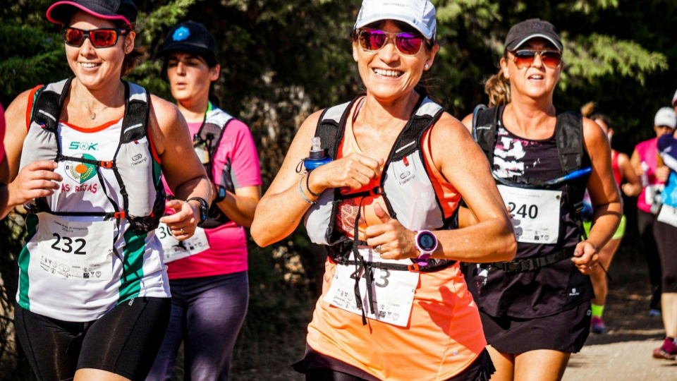 Elas usam roupas esportivas coloridas, bonés, óculos escuros e coletes de hidratação presos ao corpo. Cada uma está com um número de identificação fixado na parte frontal da roupa. A mulher em destaque, no centro da imagem, sorri enquanto corre e usa relógio esportivo no pulso.