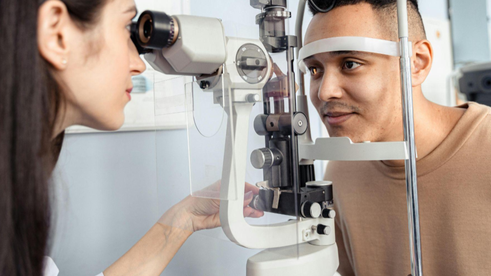 A imagem mostra um homem realizando um exame oftalmológico em um consultório. Ele está posicionado com o queixo e a testa apoiados em um equipamento oftalmológico, enquanto olha fixamente para frente.