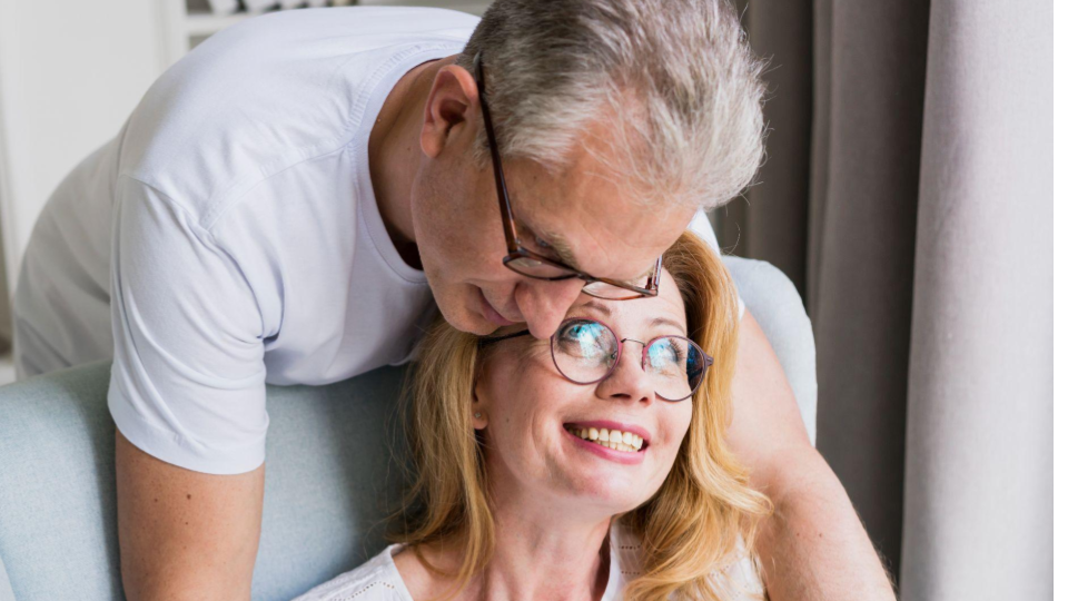A última imagem mostra um casal de meia-idade num momento de carinho e proximidade. O homem, com cabelos grisalhos e óculos de armação escura, está posicionado atrás da mulher, inclinando a cabeça suavemente contra a dela.