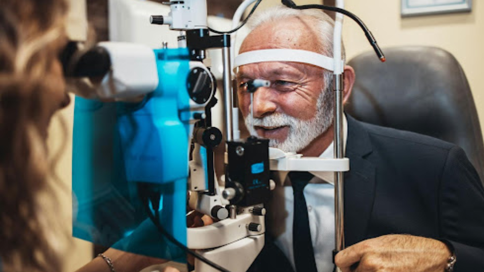A imagem mostra um homem idoso sentado em uma cadeira durante um exame oftalmológico. Ele tem cabelos brancos e barba bem aparada, e está vestido de forma elegante, com terno escuro, camisa e gravata.