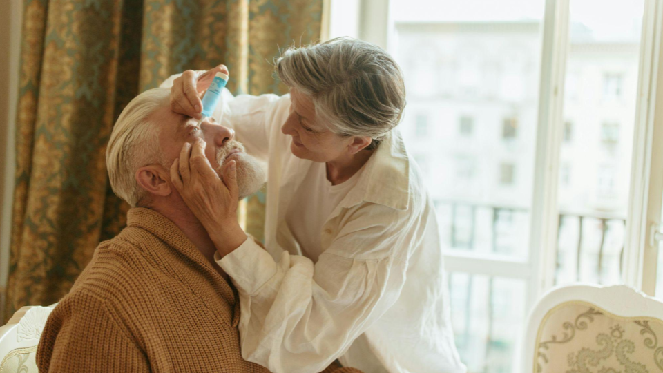Esta é uma imagem acolhedora e íntima que captura um momento de cuidado entre um casal de idosos em um ambiente doméstico iluminado.
