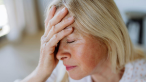 A imagem mostra o close-up de uma mulher, aparentemente de meia-idade, com cabelos loiros curtos, em um ambiente interno iluminado por luz natural. Ela está com os olhos fechados e a mão apoiada na testa, como se estivesse com dor de cabeça, preocupada, cansada ou sob estresse emocional.