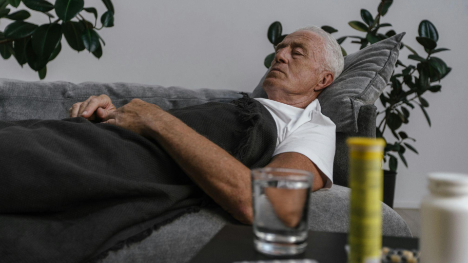 A imagem mostra um homem idoso deitado em um sofá, aparentemente descansando ou dormindo, coberto por uma manta escura. Ele está com a cabeça apoiada em um travesseiro e os olhos fechados, sugerindo repouso ou recuperação.