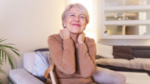 A imagem mostra uma mulher idosa sentada em uma poltrona confortável, dentro de uma sala de estar bem iluminada e aconchegante. Ela tem cabelos curtos e grisalhos e veste uma blusa de manga longa em tom neutro (marrom claro).