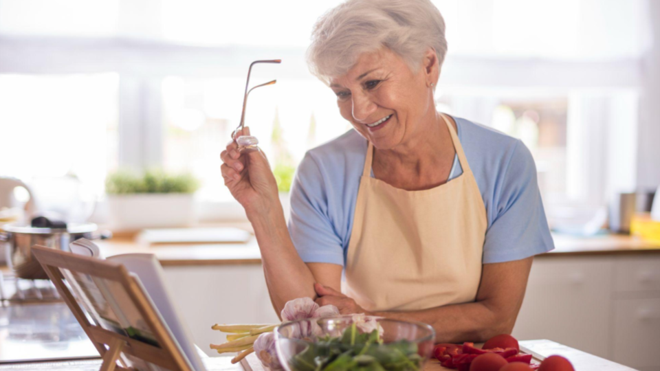 A imagem mostra uma mulher idosa em uma cozinha clara e aconchegante, preparando alimentos enquanto interage com um dispositivo digital.