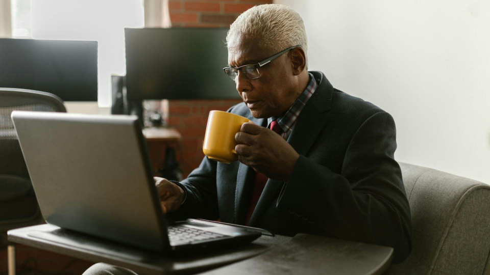 A imagem mostra um homem idoso em um ambiente de trabalho moderno, sentado diante de um notebook enquanto toma uma bebida quente.