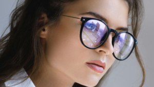 A imagem mostra um close-up do rosto de uma mulher usando óculos com lentes grandes e armação escura. Ela está levemente de perfil, olhando para frente com expressão séria e concentrada.