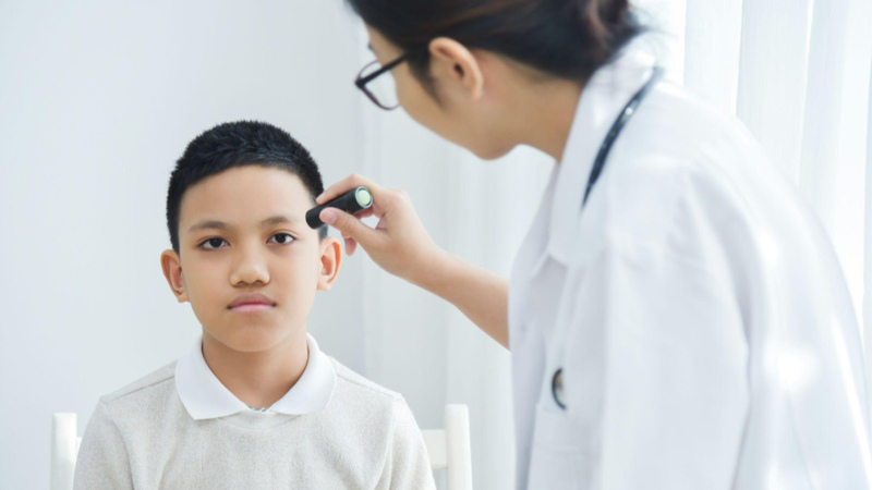 Um menino está sentado e olhando para a frente enquanto uma médica, usando jaleco branco e óculos, realiza um exame.