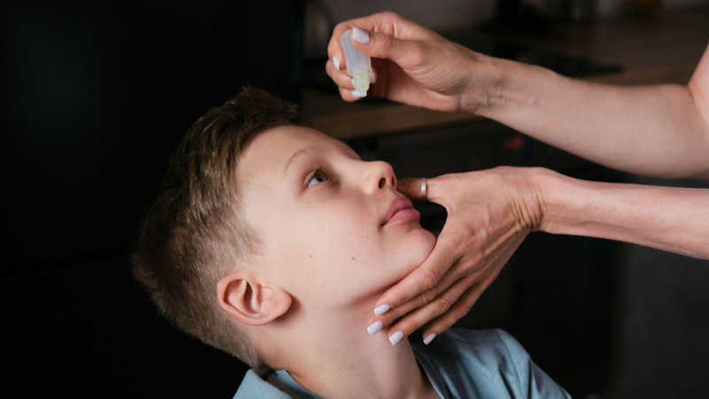 Uma imagem em close-back que mostra um menino com o rosto inclinado para trás enquanto recebe gotas de um colírio ou medicamento nos olhos.