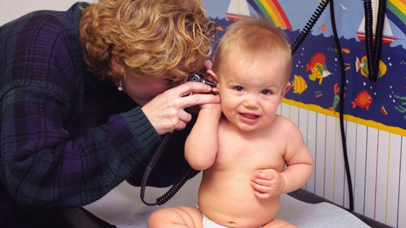 Uma médica pediatra examina o ouvido de um bebê sorridente usando um otoscópio. O bebê está sentado em uma mesa de exame em um consultório decorado com temas infantis (desenhos de barcos e arco-íris ao fundo).