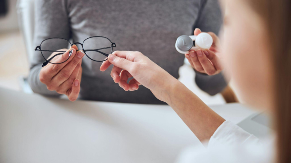 Uma pessoa entrega um par de óculos para outra durante uma consulta oftalmológica. Em uma das mãos, há um estojo com lentes de contato, sugerindo uma conversa sobre opções de correção visual. A cena transmite atendimento especializado, escolha de soluções para a visão e cuidados com a saúde ocular.